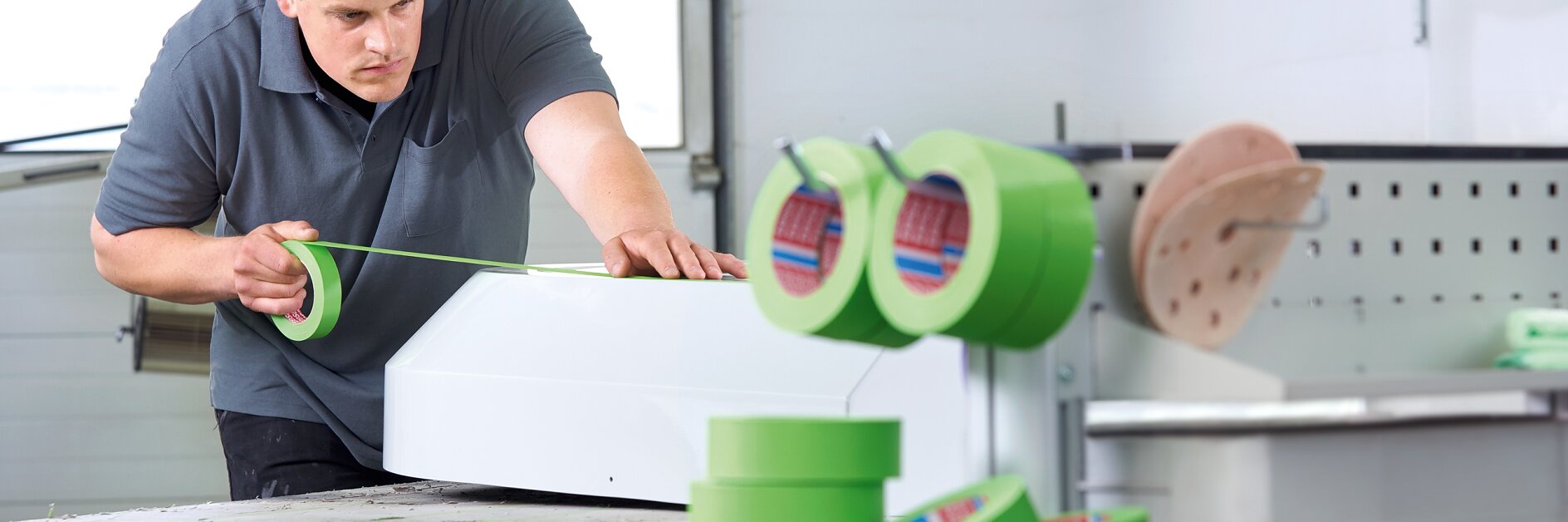 A man in a workshop applies green tesa tape to a white object on a wooden workbench. Rolls of green tesa tape are visible in the foreground. He is wearing a gray shirt and appears focused on the task. The background shows a workshop setting with tools and equipment. (This text has been generated by AI)