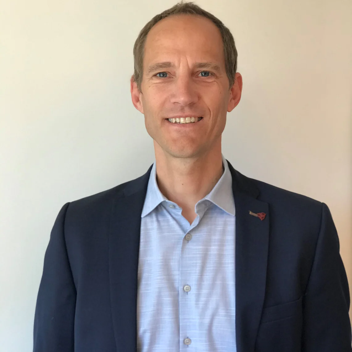 A person with short hair is wearing a navy blazer over a light blue button-up shirt. They are standing in front of a plain off-white wall, smiling slightly at the camera. (This text has been generated by AI)