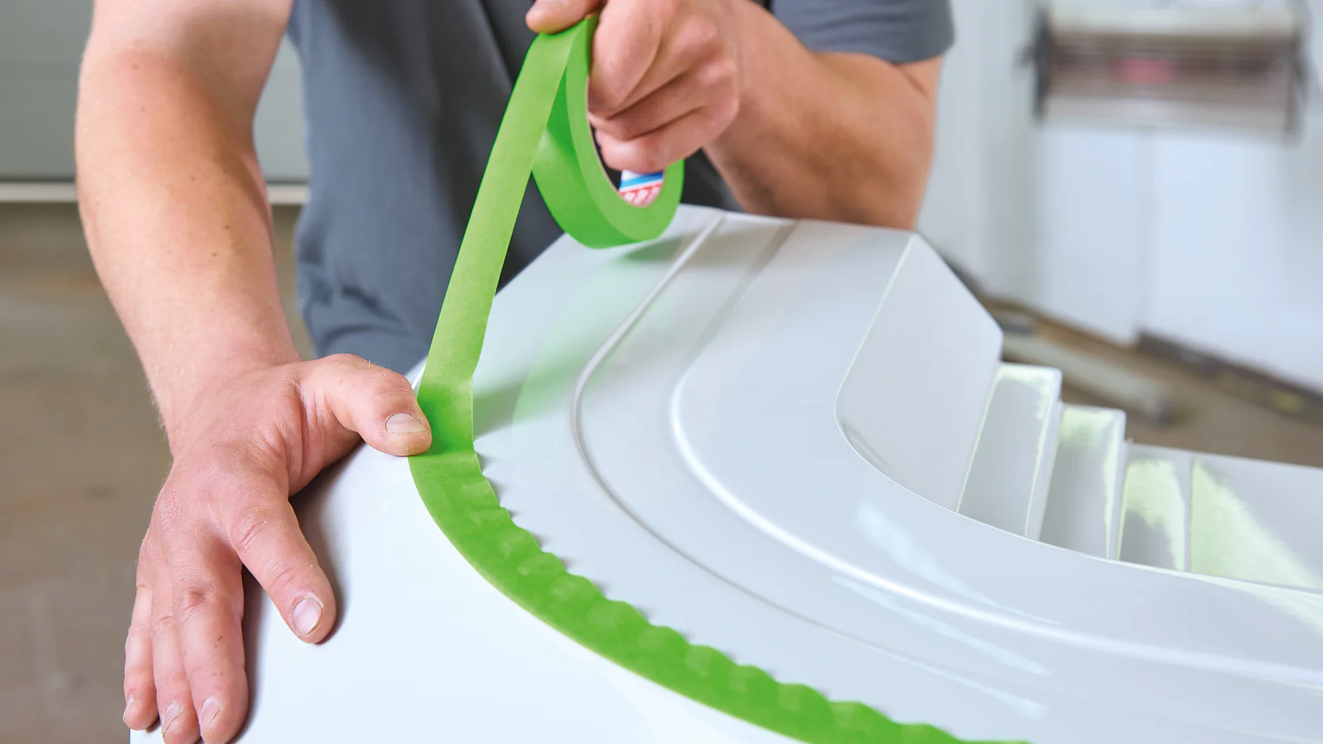 A person is applying green tesa tape to the edge of a white surface, using both hands to carefully align it. The individual is wearing a gray shirt, fully focused on the tesa tape application process. (This text has been generated by AI)