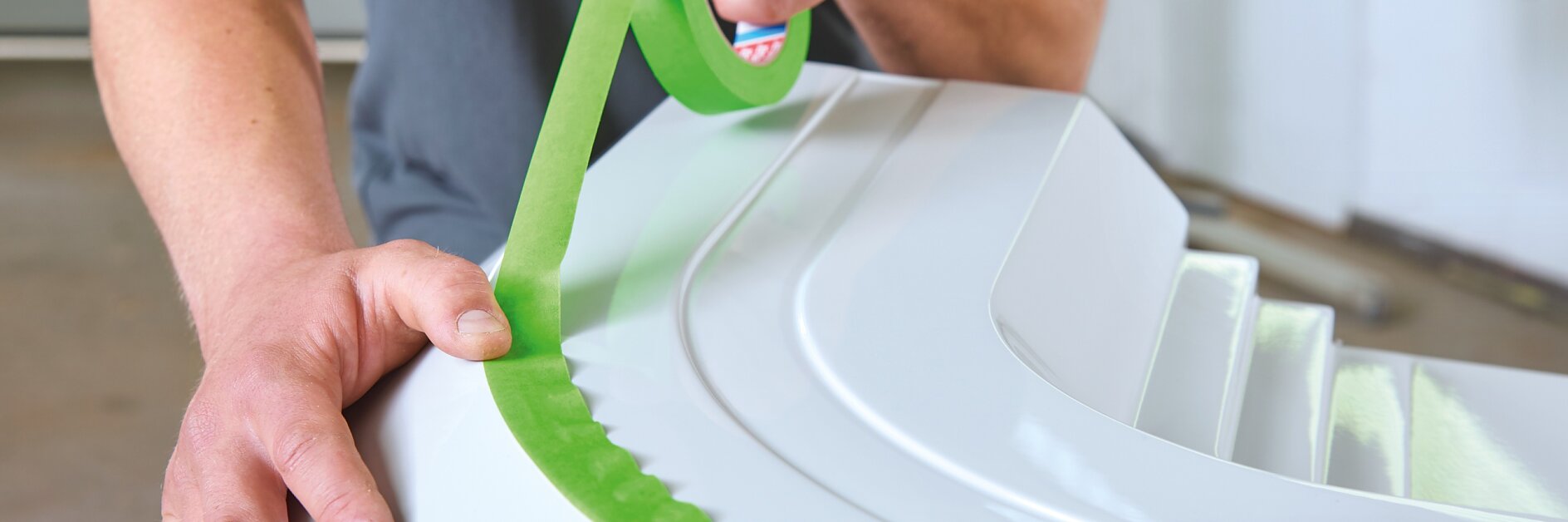 A person is applying green tesa tape to the edge of a white surface, using both hands to carefully align it. The individual is wearing a gray shirt, fully focused on the tesa tape application process. (This text has been generated by AI)