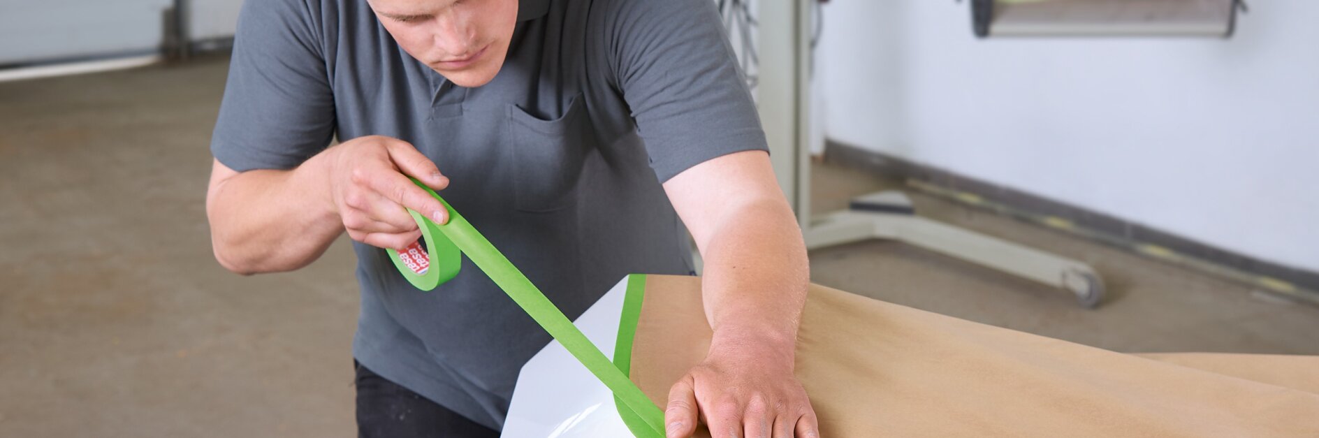 A man in a gray t-shirt is carefully applying green tesa tape to the edge of a piece of brown paper covering a section of a white car. The scene takes place in a garage or workshop setting. (This text has been generated by AI)