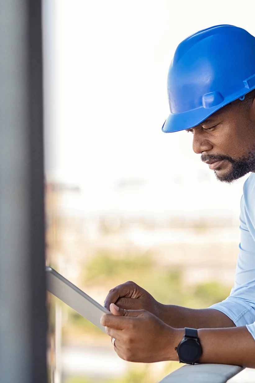 Reference building, Raiffeisenbank Lugano (Tessin), Switzerland, Photo: Daniel Sumesgutner A construction worker on an upper floor of a building site, wearing a blue helmet, leaning on the scaffolding with one arm and holding a paper that he is inspecting, with possible construction tapes visible in the background. Photo: Daniel Sumesgutner