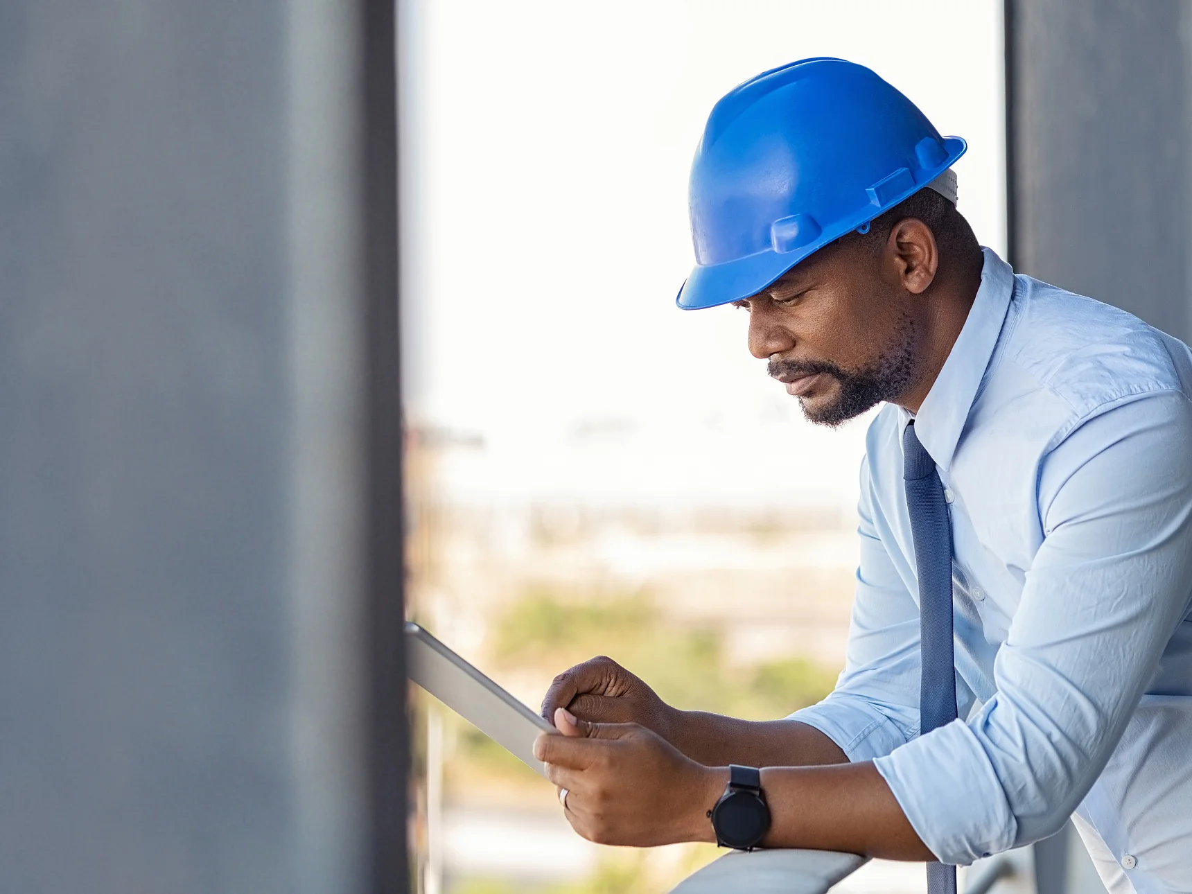 Reference building, Raiffeisenbank Lugano (Tessin), Switzerland, Photo: Daniel Sumesgutner A construction worker on an upper floor of a building site, wearing a blue helmet, leaning on the scaffolding with one arm and holding a paper that he is inspecting, with possible construction tapes visible in the background. Photo: Daniel Sumesgutner