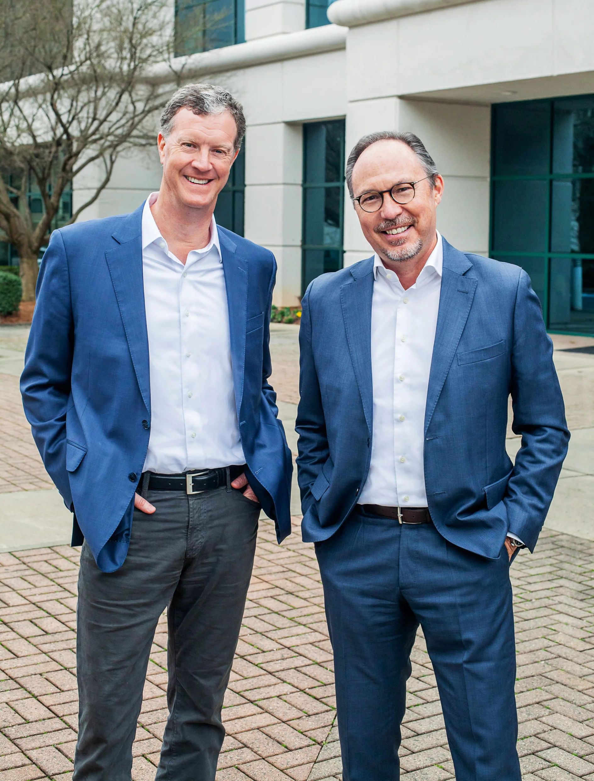 Paul Sullivan and Daniel Germain Two men in suits stand outdoors on a paved area. Both are wearing blue blazers, white shirts, and have their hands in their pockets. The man on the left is wearing gray pants, while the man on the right is wearing blue pants. A building and trees are in the background. (This text has been generated by AI)