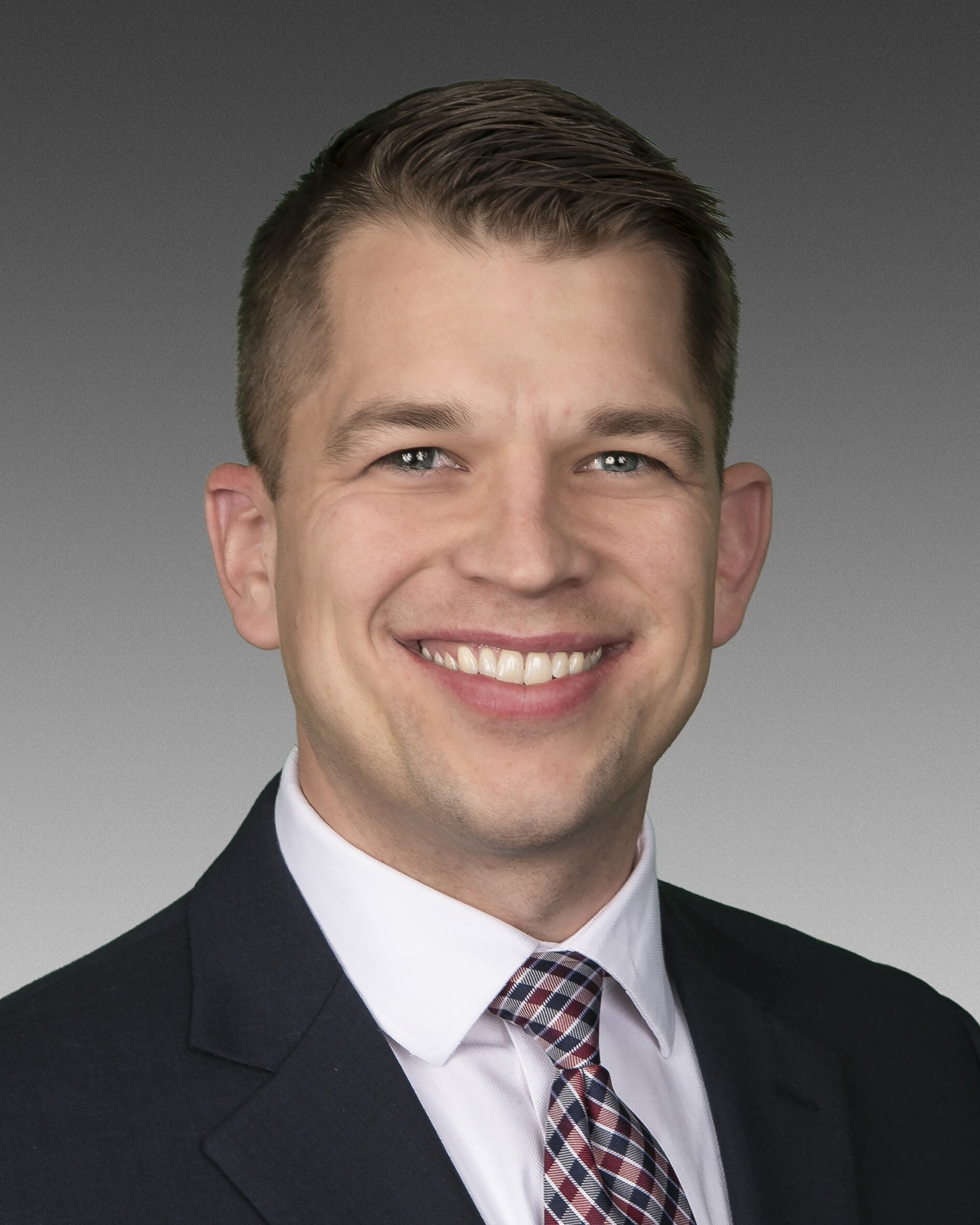 A person with short hair, wearing a dark suit, white shirt, and checkered tie, is smiling in a portrait against a gradient gray background. (This text has been generated by AI)