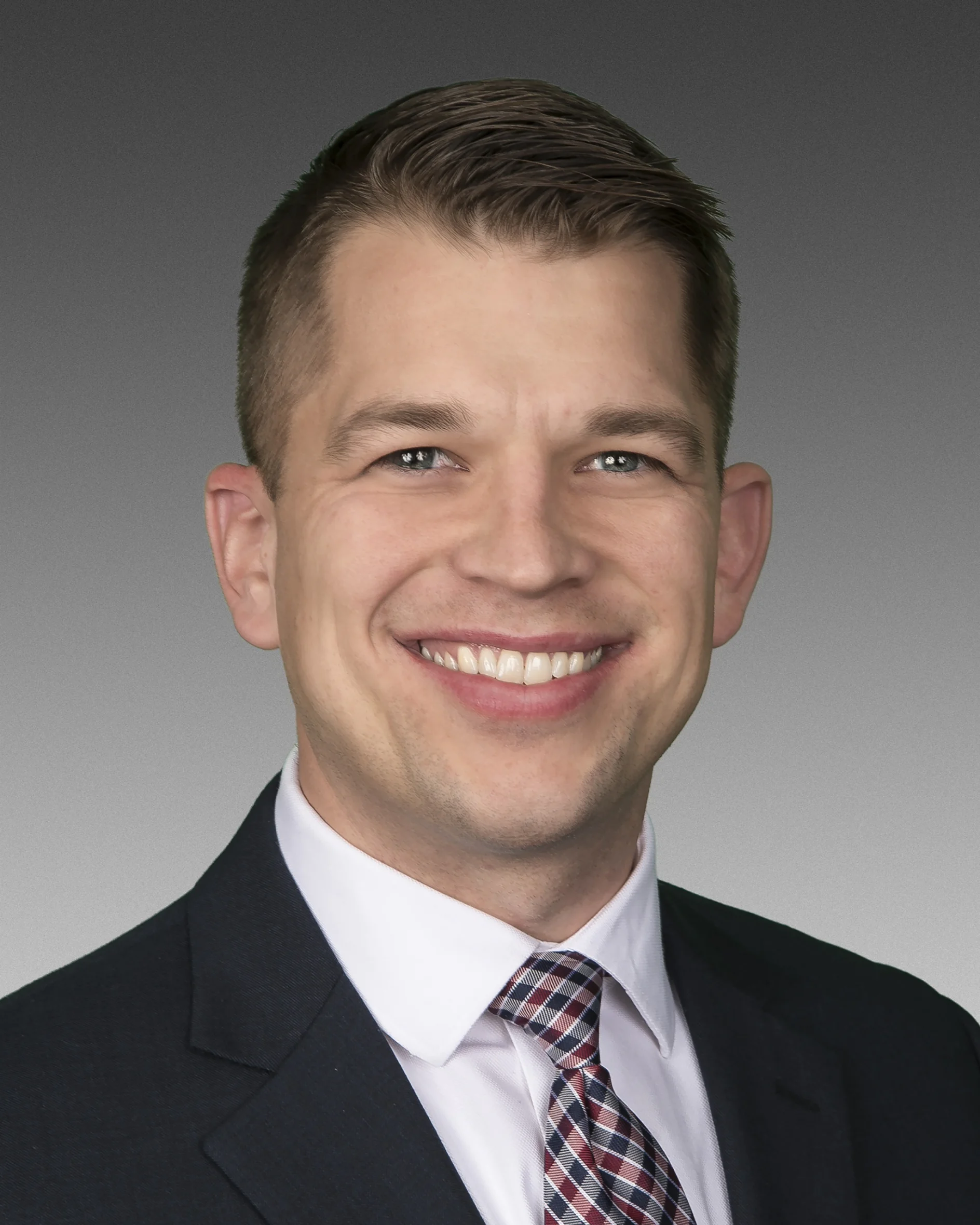 A person with short hair, wearing a dark suit, white shirt, and checkered tie, is smiling in a portrait against a gradient gray background. (This text has been generated by AI)