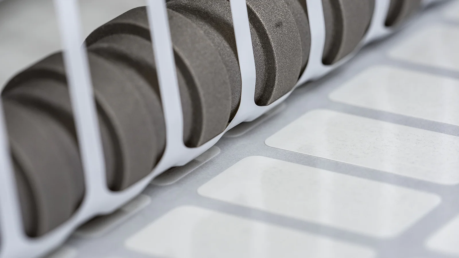 Close-up of a set of black gears positioned next to a row of white labels running along a conveyor system, partially enclosed by white vertical bars. The scene suggests an industrial setting, likely related to printing or labeling using tesa tape. (This text has been generated by AI)