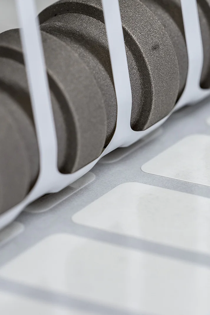 Close-up of a set of black gears positioned next to a row of white labels running along a conveyor system, partially enclosed by white vertical bars. The scene suggests an industrial setting, likely related to printing or labeling using tesa tape. (This text has been generated by AI)