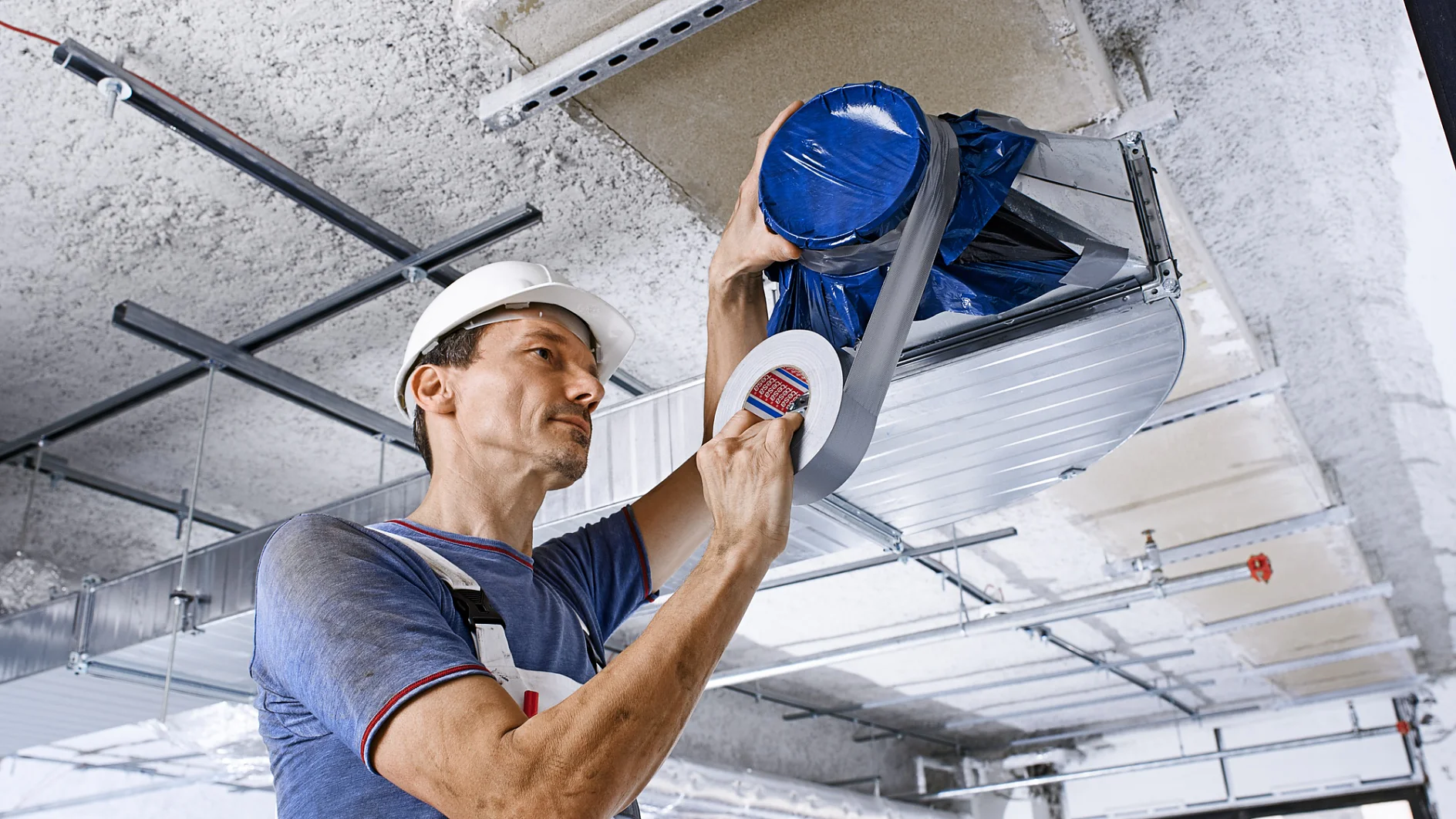 A craftsman standing close to an unfinished ceiling is holding a roll of tesa cloth tape and using it to work on a pipe attached to the ceiling. The entire unfinished ceiling is visible.