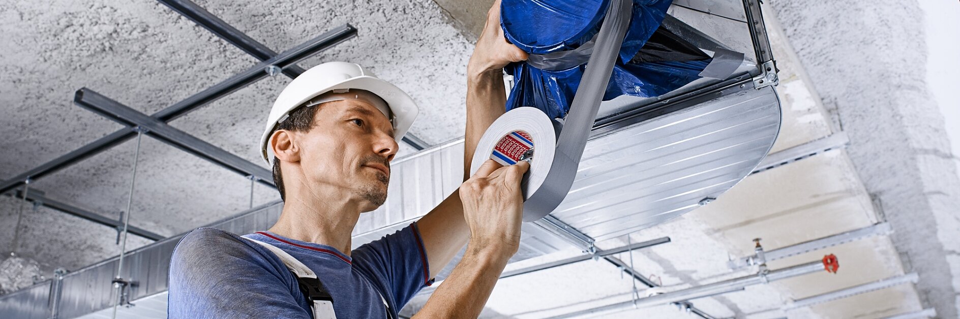 A craftsman standing close to an unfinished ceiling is holding a roll of tesa cloth tape and using it to work on a pipe attached to the ceiling. The entire unfinished ceiling is visible.