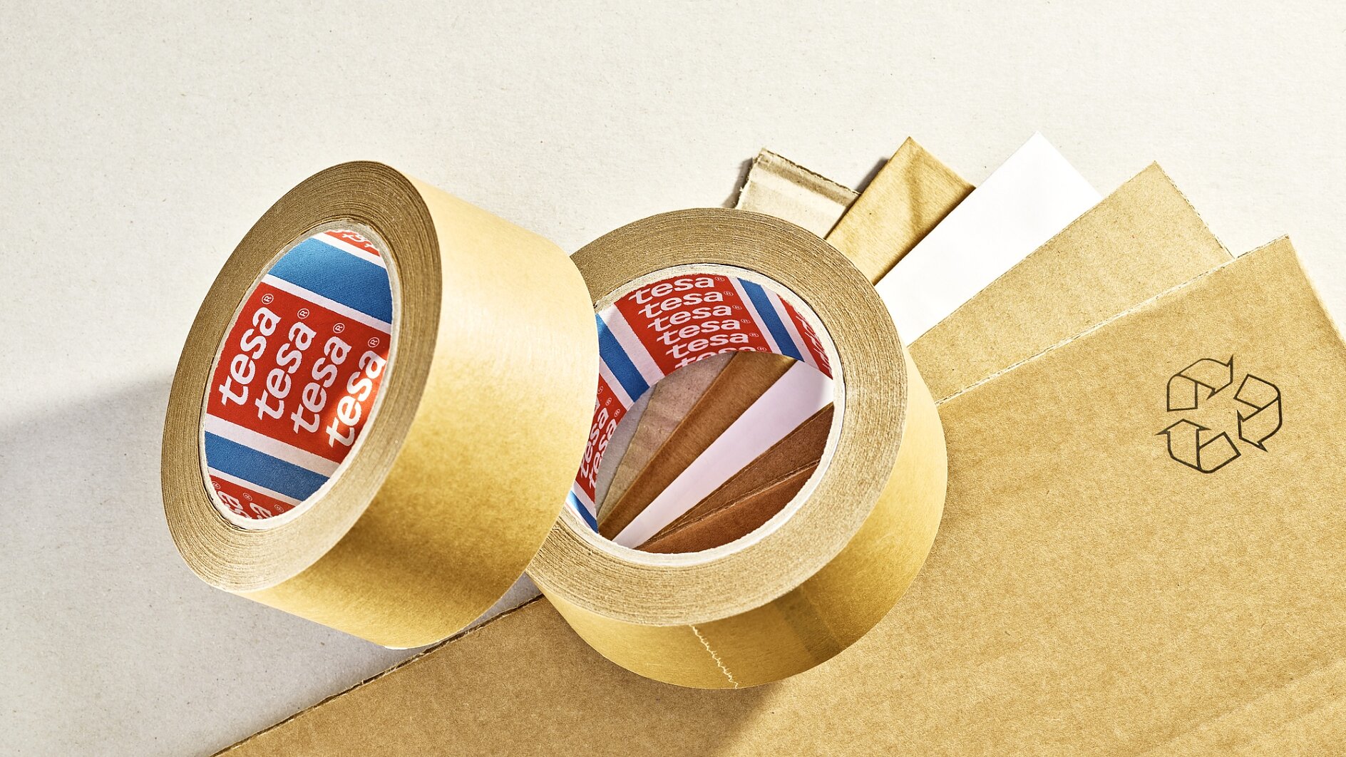 Two rolls of brown adhesive tesa tape are placed on sheets of brown cardboard. The tesa tape has red and blue labels with branding. A recycling symbol is printed on one of the cardboard sheets. (This text has been generated by AI)