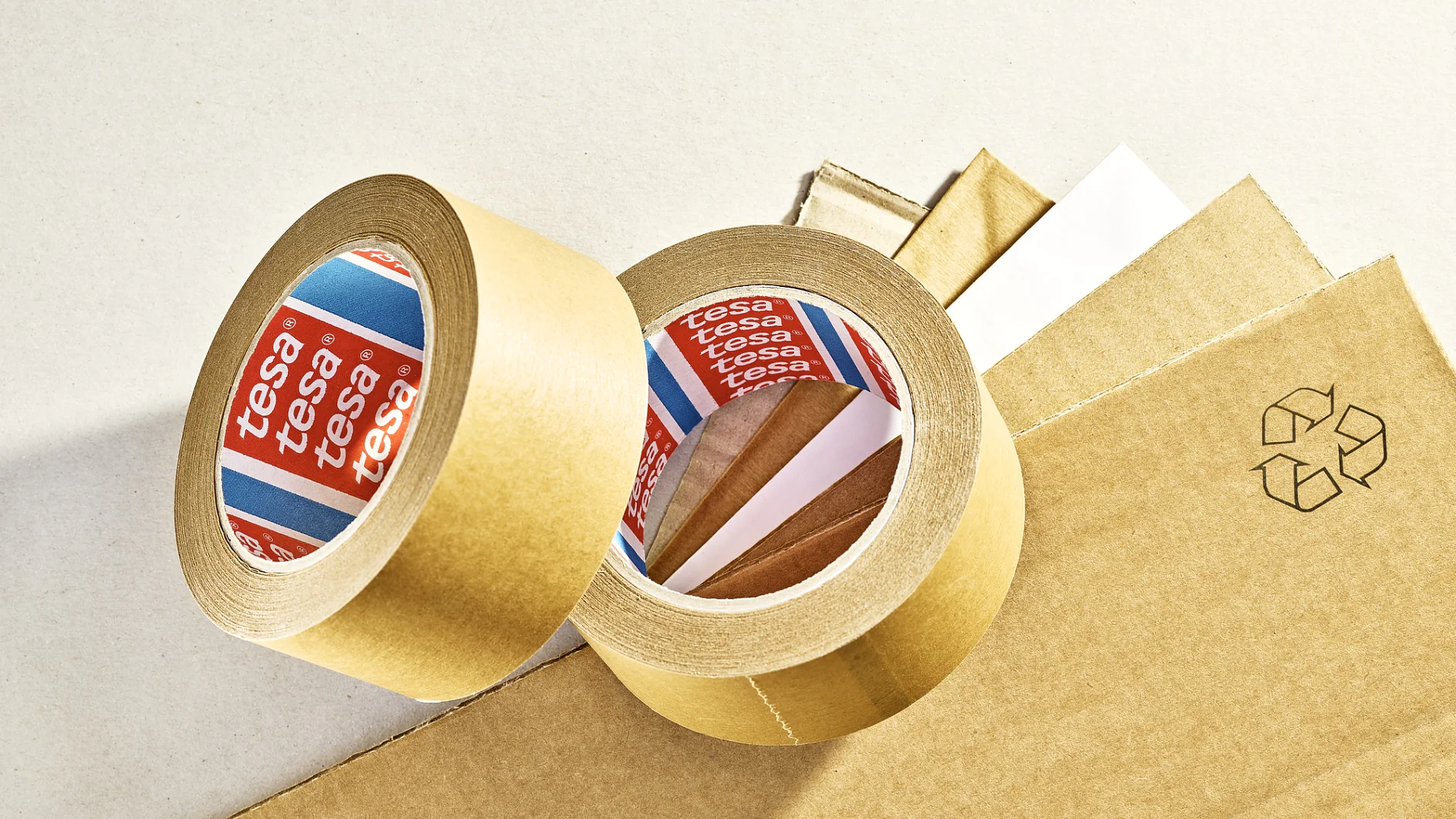 Two rolls of brown adhesive tesa tape are placed on sheets of brown cardboard. The tesa tape has red and blue labels with branding. A recycling symbol is printed on one of the cardboard sheets. (This text has been generated by AI)