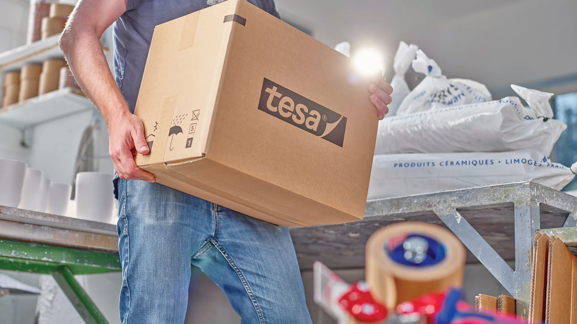 A person holding a large cardboard box labeled tesa in a workshop setting. In the foreground, theres a table with tesa tape and a dispenser. Bags of materials are stacked in the background. The person is wearing a dark t-shirt and jeans. (This text has been generated by AI)