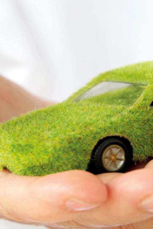 A hand is holding a small model car covered in grass-like material. The car appears to be green and eco-friendly, symbolizing environmental sustainability. The background is blurry and white. (This text has been generated by AI)