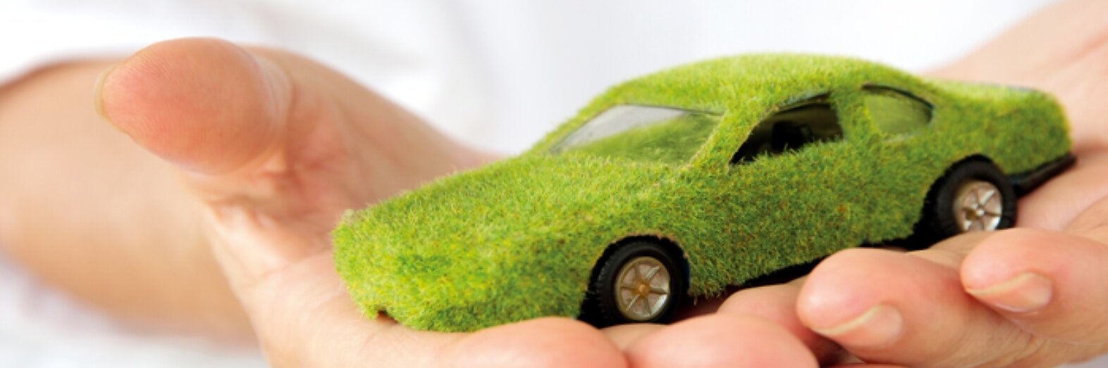 A hand is holding a small model car covered in grass-like material. The car appears to be green and eco-friendly, symbolizing environmental sustainability. The background is blurry and white. (This text has been generated by AI)