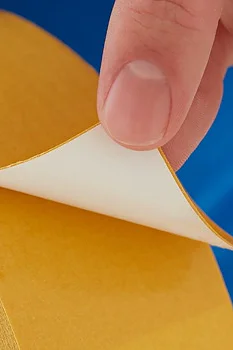A hand is peeling back the protective paper from a yellow double-sided tesa tape, revealing the adhesive side. The person is wearing a blue and red garment. (This text has been generated by AI)