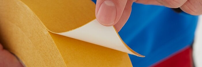 A hand is peeling back the protective paper from a yellow double-sided tesa tape, revealing the adhesive side. The person is wearing a blue and red garment. (This text has been generated by AI)