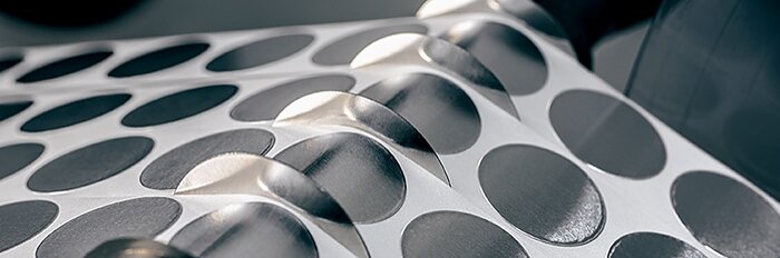 A sheet of round, metallic stickers is being processed through a machine. The stickers are arranged in neat rows on the sheet, and the machine appears to be part of a laminating or stamping process using tesa tape. (This text has been generated by AI)