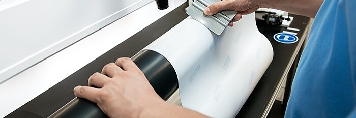 A person applies a sheet of white material onto a cylindrical surface using a squeegee-like tool. The process appears to be part of an industrial or printing setup, with tesa tape machinery visible in the background. (This text has been generated by AI)