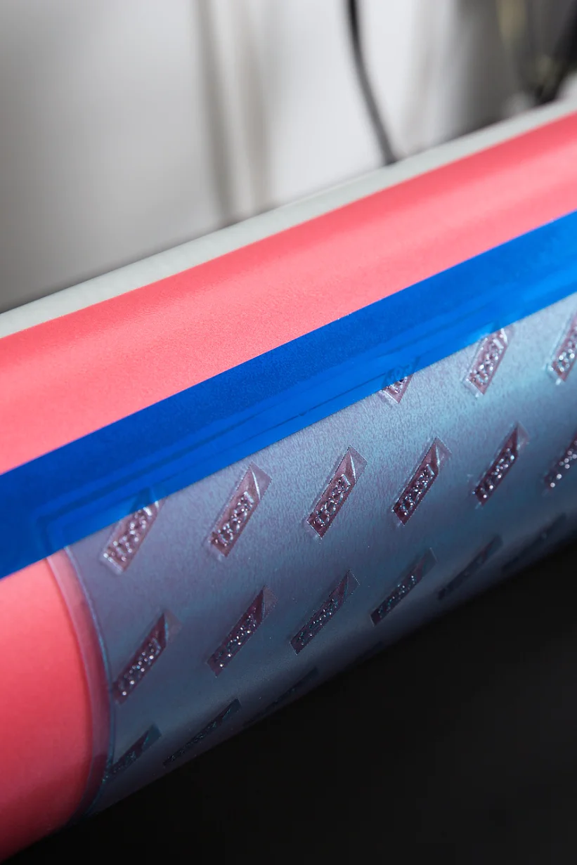 A close-up of a cylindrical object, possibly part of a machine. The object is pink with a strip of silver material featuring a pattern of raised shapes, secured with blue tesa tape. The background is out of focus. (This text has been generated by AI)