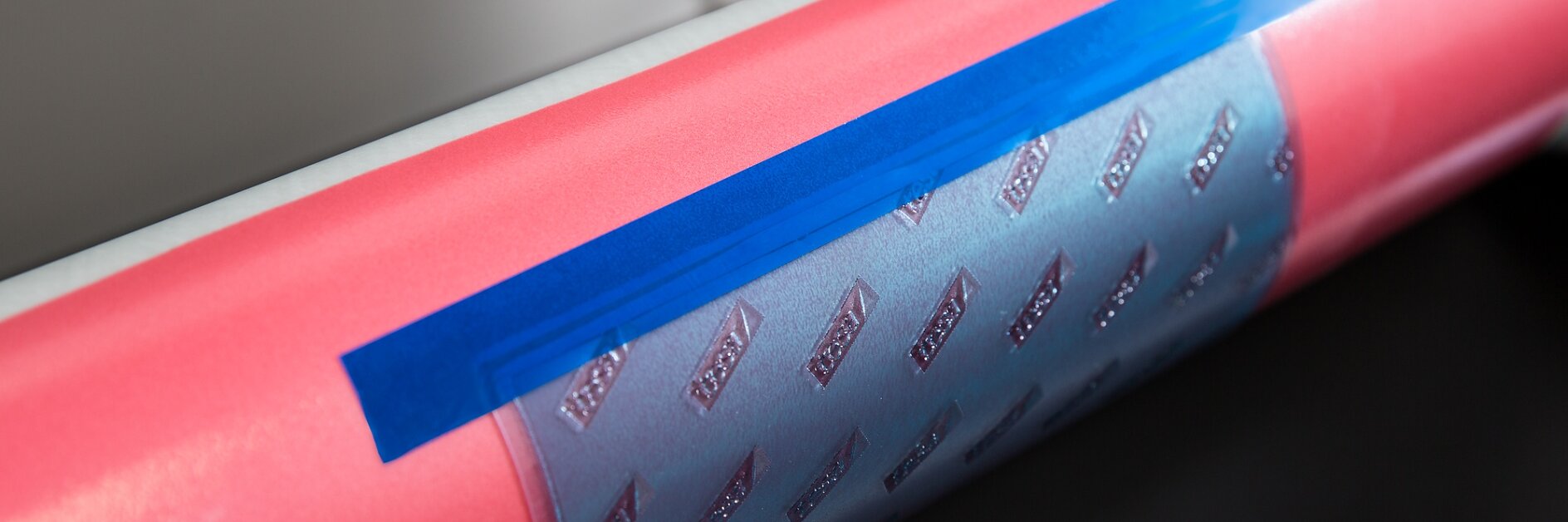 A close-up of a cylindrical object, possibly part of a machine. The object is pink with a strip of silver material featuring a pattern of raised shapes, secured with blue tesa tape. The background is out of focus. (This text has been generated by AI)