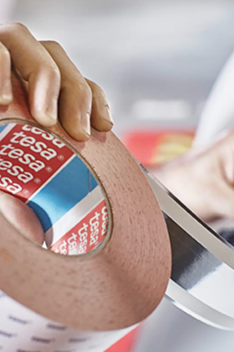 A person holds a large roll of adhesive tesa tape with a red and blue inner label reading tesa. Another hand is cutting a piece of the tesa tape with a dark-colored tool. The background is blurred, emphasizing the focus on the tesa tape and hands. (This text has been generated by AI)