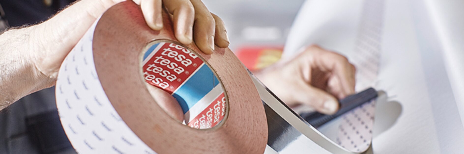 A person holds a large roll of adhesive tesa tape with a red and blue inner label reading tesa. Another hand is cutting a piece of the tesa tape with a dark-colored tool. The background is blurred, emphasizing the focus on the tesa tape and hands. (This text has been generated by AI)