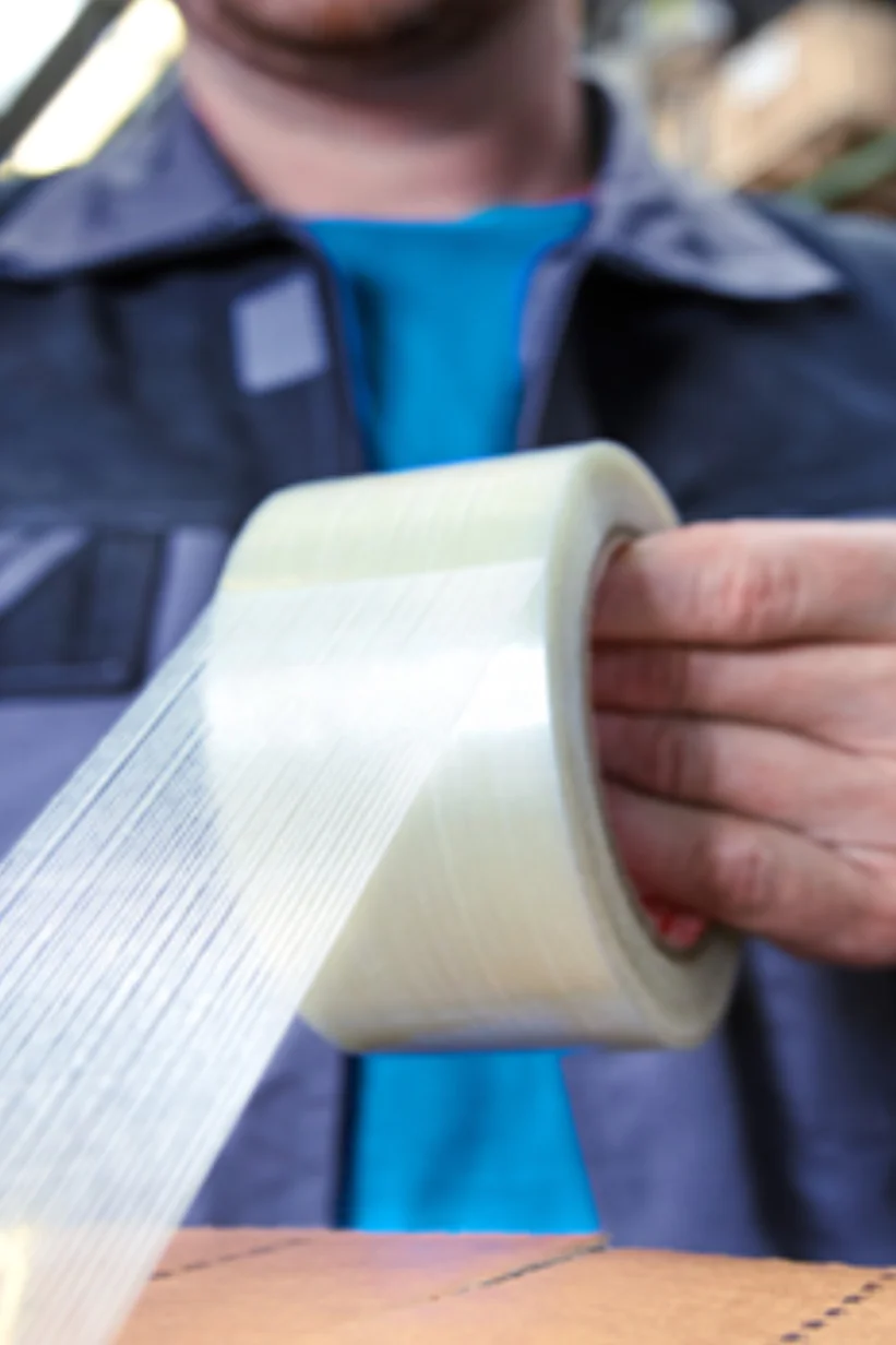 A person wearing a blue shirt and a vest is sealing a cardboard box with transparent packing tesa tape. They are in a warehouse or storage setting, surrounded by shelves filled with various items. (This text has been generated by AI)