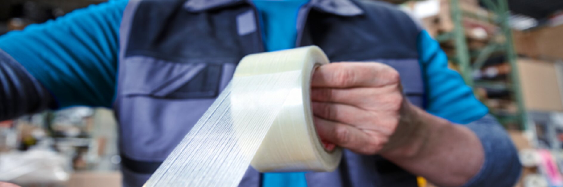 A person wearing a blue shirt and a vest is sealing a cardboard box with transparent packing tesa tape. They are in a warehouse or storage setting, surrounded by shelves filled with various items. (This text has been generated by AI)