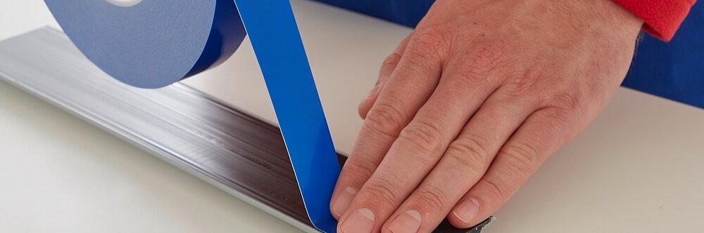A person wearing a red sleeve applies blue adhesive tesa tape to a narrow, flat metal strip on a white surface. The hand is holding and pressing down the tesa tape for a smooth application. (This text has been generated by AI)