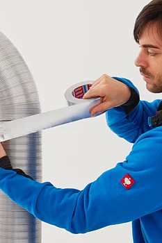 A person in a blue jacket is applying tesa tape to seal the joint of a large, curved air duct. A tool box is visible on a table beside them. The background is plain and white. (This text has been generated by AI)