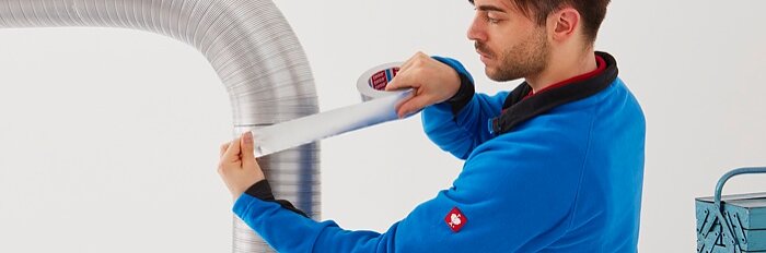 A person in a blue jacket is applying tesa tape to seal the joint of a large, curved air duct. A tool box is visible on a table beside them. The background is plain and white. (This text has been generated by AI)