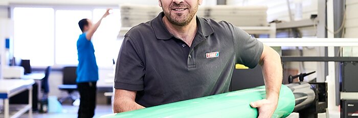 A man in a gray polo shirt holds a large roll of green tesa tape in a well-lit workspace. Another person wearing a blue shirt is blurred in the background. Shelves and equipment are visible in the room. (This text has been generated by AI)