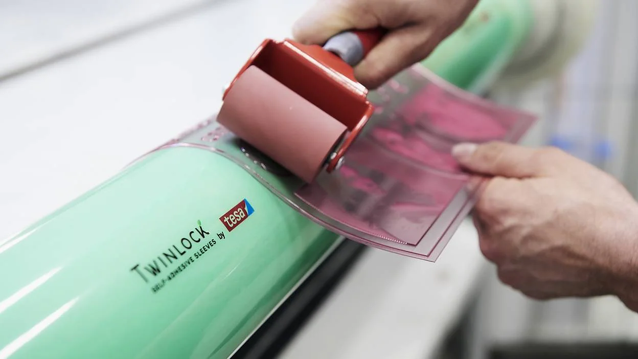 twinlock-by-tesa-plate-mounting,674977_crop16x9_12 A person uses a red roller to apply a pink adhesive sheet labeled tesa tape TWINLOCK to a green cylindrical surface. The background is blurred, focusing on the hands and tools. (This text has been generated by AI)