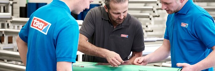 Three men are examining a green cylindrical object in an industrial setting. Two men wear blue shirts with a red and white logo, while the third man wears a gray shirt with the same logo. Metal bars are visible in the background, and tesa tape is being used on various components of the object. (This text has been generated by AI)