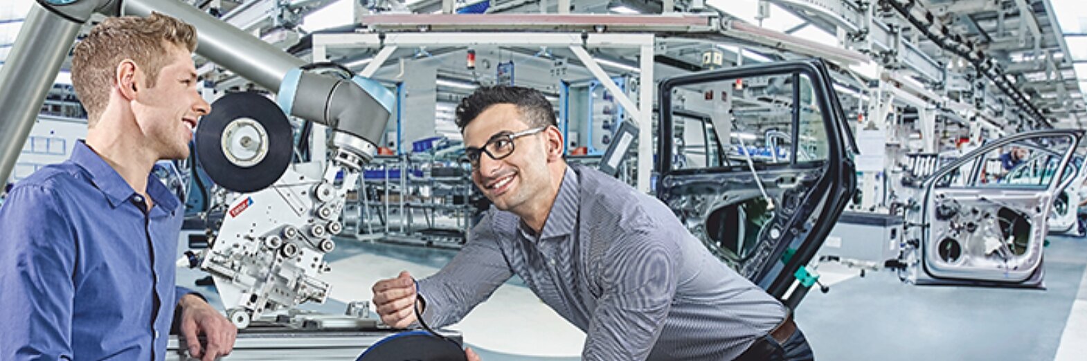 Two men are interacting in an industrial setting. One holds a robotic component, while the other appears to discuss or demonstrate something related, possibly involving tesa tape. Car doors and machinery are in the background, suggesting a manufacturing environment. (This text has been generated by AI)