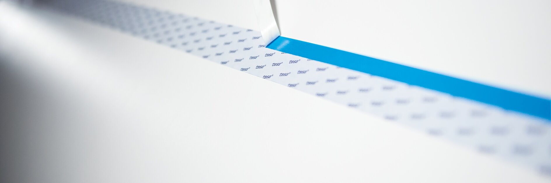 A close-up of a precision cutting tool slicing through paper, revealing a narrow blue strip of tesa tape underneath. The surface being cut appears white with a lightly dotted pattern. The scene is well-lit, emphasizing the sharpness of the cutting edge. (This text has been generated by AI)