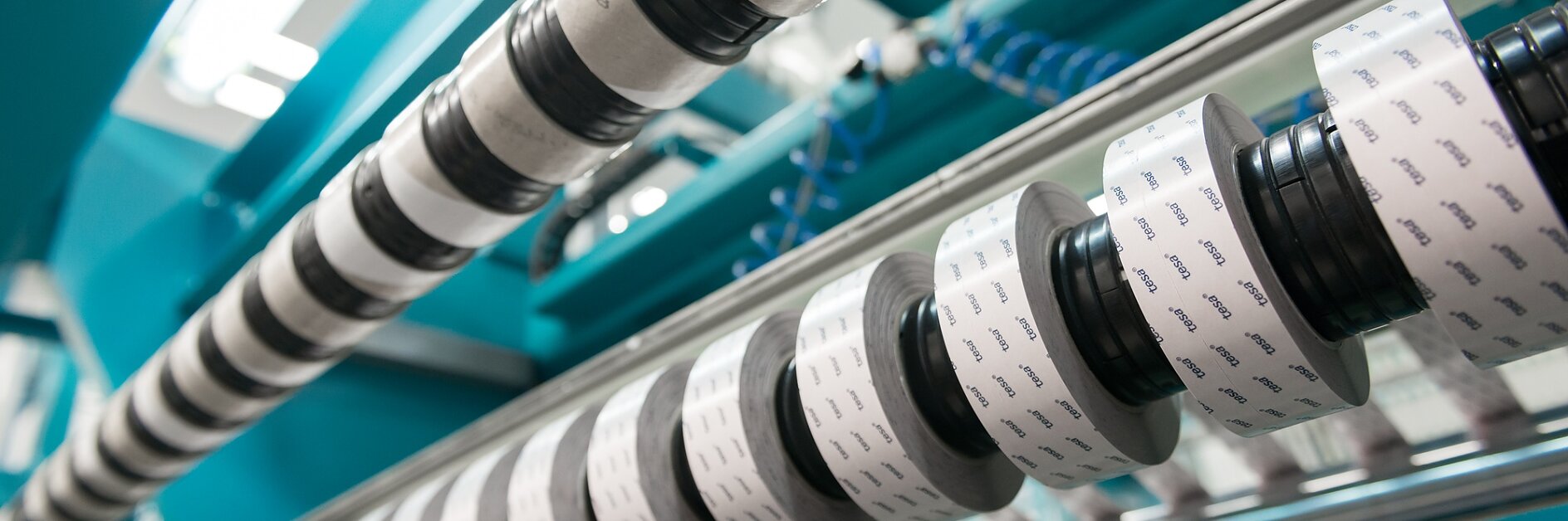 A close-up view of a machine with multiple rollers wrapped in black and white striped material and a few rolls of film with printed patterns. The machine parts are made of metal and are arranged in a row. The background is blurred, featuring tesa tape prominently used in the setup. (This text has been generated by AI)