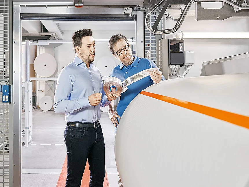 Two men are inspecting a large roll of paper in an industrial setting. One man is holding a sample strip of tesa tape and gesturing, while the other observes. The background shows additional paper rolls and industrial equipment. (This text has been generated by AI)