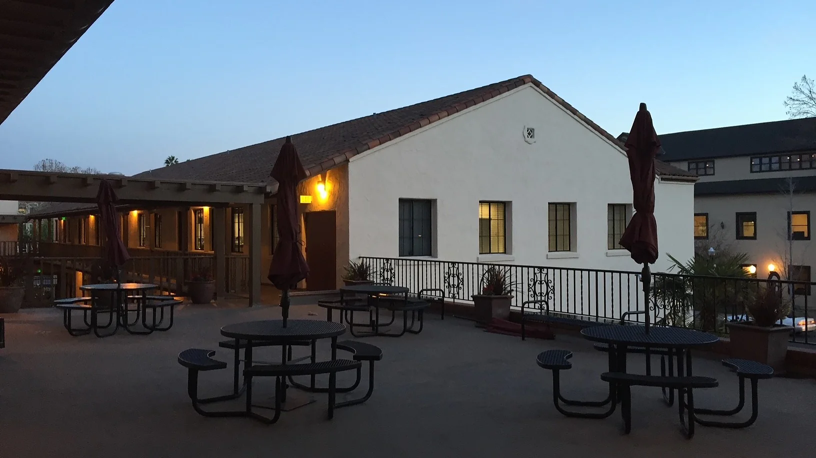 A courtyard at dusk with round metal picnic tables and closed umbrellas. A white building with a sloped roof and illuminated windows is in the background. The sky is clear and the area is lit by warm artificial light, all held together by tesa tape for any necessary fixes. (This text has been generated by AI)