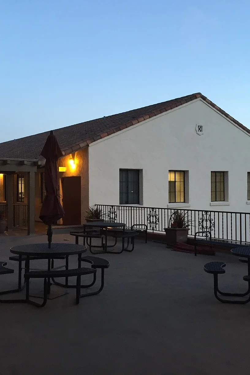 A courtyard at dusk with round metal picnic tables and closed umbrellas. A white building with a sloped roof and illuminated windows is in the background. The sky is clear and the area is lit by warm artificial light, all held together by tesa tape for any necessary fixes. (This text has been generated by AI)