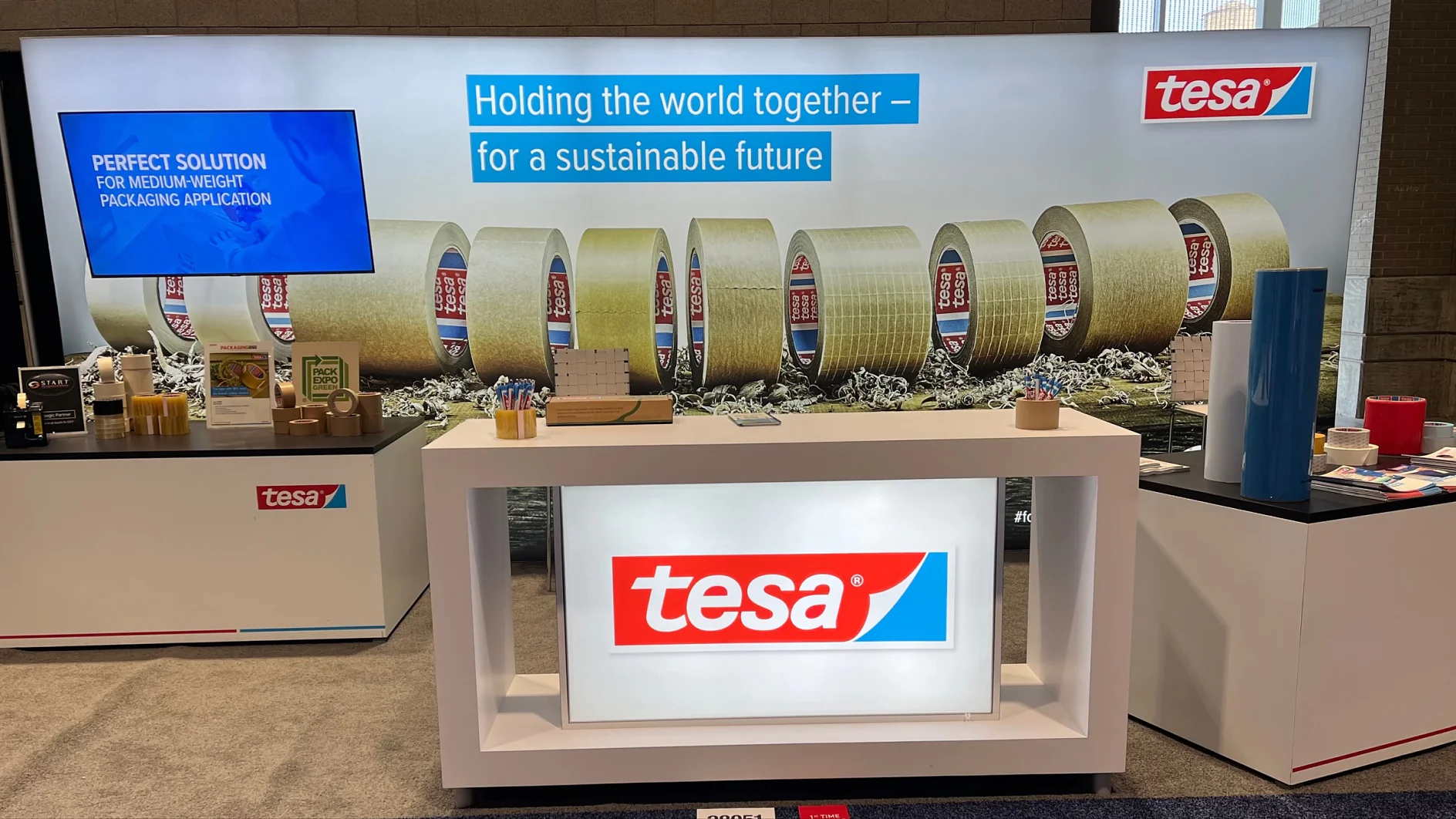 Exhibition booth showcasing tesa products, featuring a lit display with the tesa logo. The backdrop displays rows of tesa tape rolls. A screen on the left shows text about packaging applications. Brochures and samples are arranged on the counters. (This text has been generated by AI)