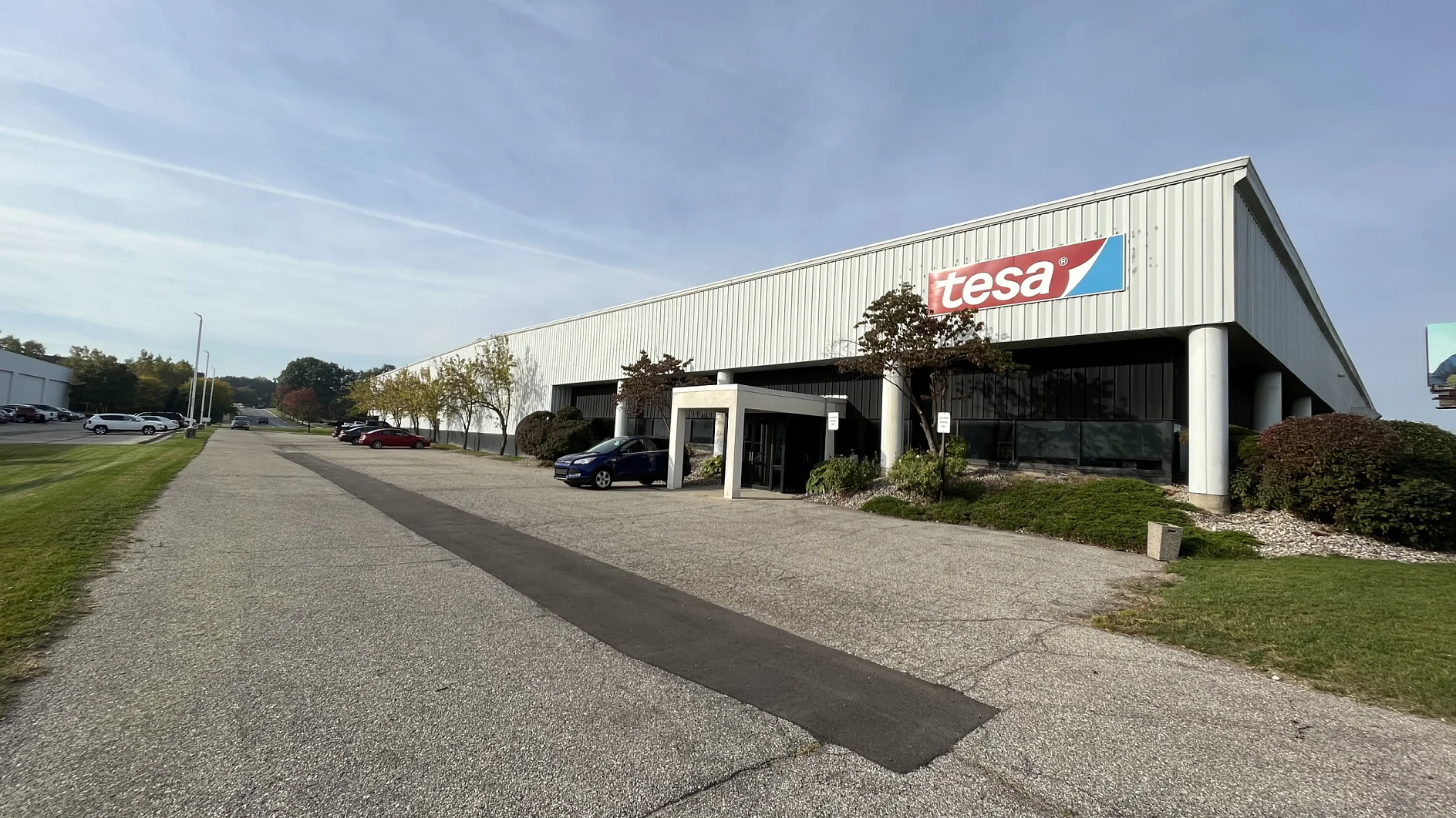 A large industrial building with a sign reading tesa on its facade. The structure has a row of windows and a covered entryway. A road runs alongside, with trees and shrubs lining the lawn. The sky is partly cloudy. (This text has been generated by AI)