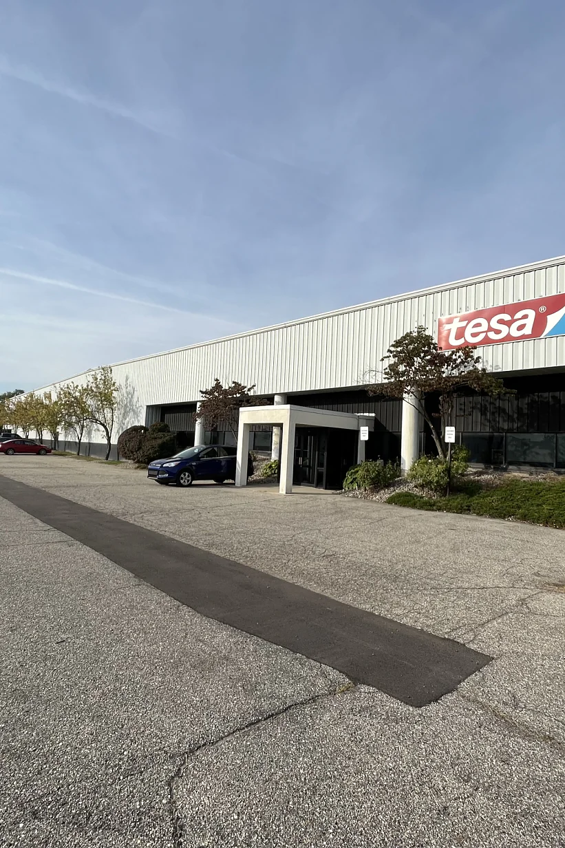 A large industrial building with a sign reading tesa on its facade. The structure has a row of windows and a covered entryway. A road runs alongside, with trees and shrubs lining the lawn. The sky is partly cloudy. (This text has been generated by AI)