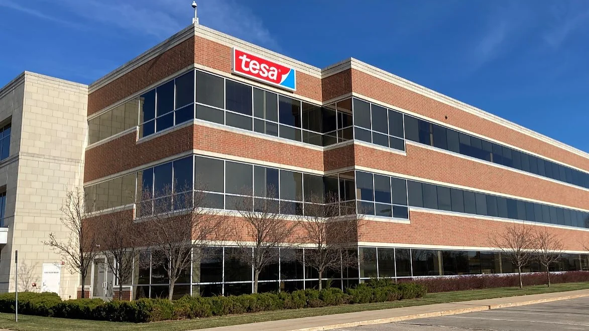 SIGNAGE 2 CROPPED 2 A three-story office building features a red and white tesa logo on the top corner. The building has large glass windows and is surrounded by a parking area with a few trees and shrubs. The sky is clear and blue. (This text has been generated by AI)