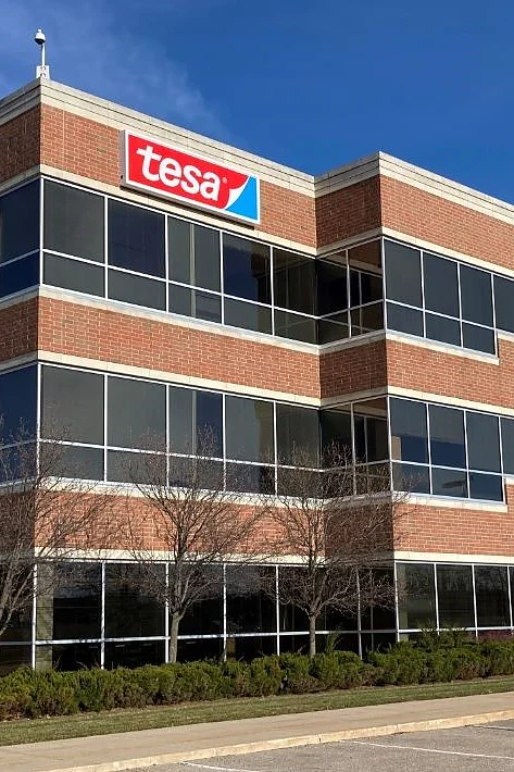 A three-story office building features a red and white tesa logo on the top corner. The building has large glass windows and is surrounded by a parking area with a few trees and shrubs. The sky is clear and blue. (This text has been generated by AI)