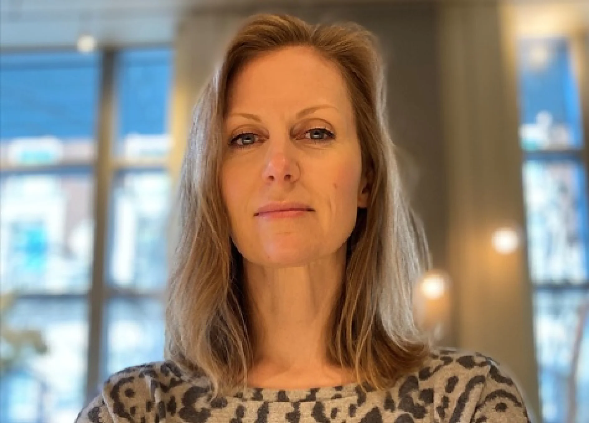 A person with shoulder-length hair and a neutral expression stands indoors, wearing a leopard print top. They are in front of large windows, through which a blurred cityscape is visible. The lighting is warm and soft. (This text has been generated by AI)