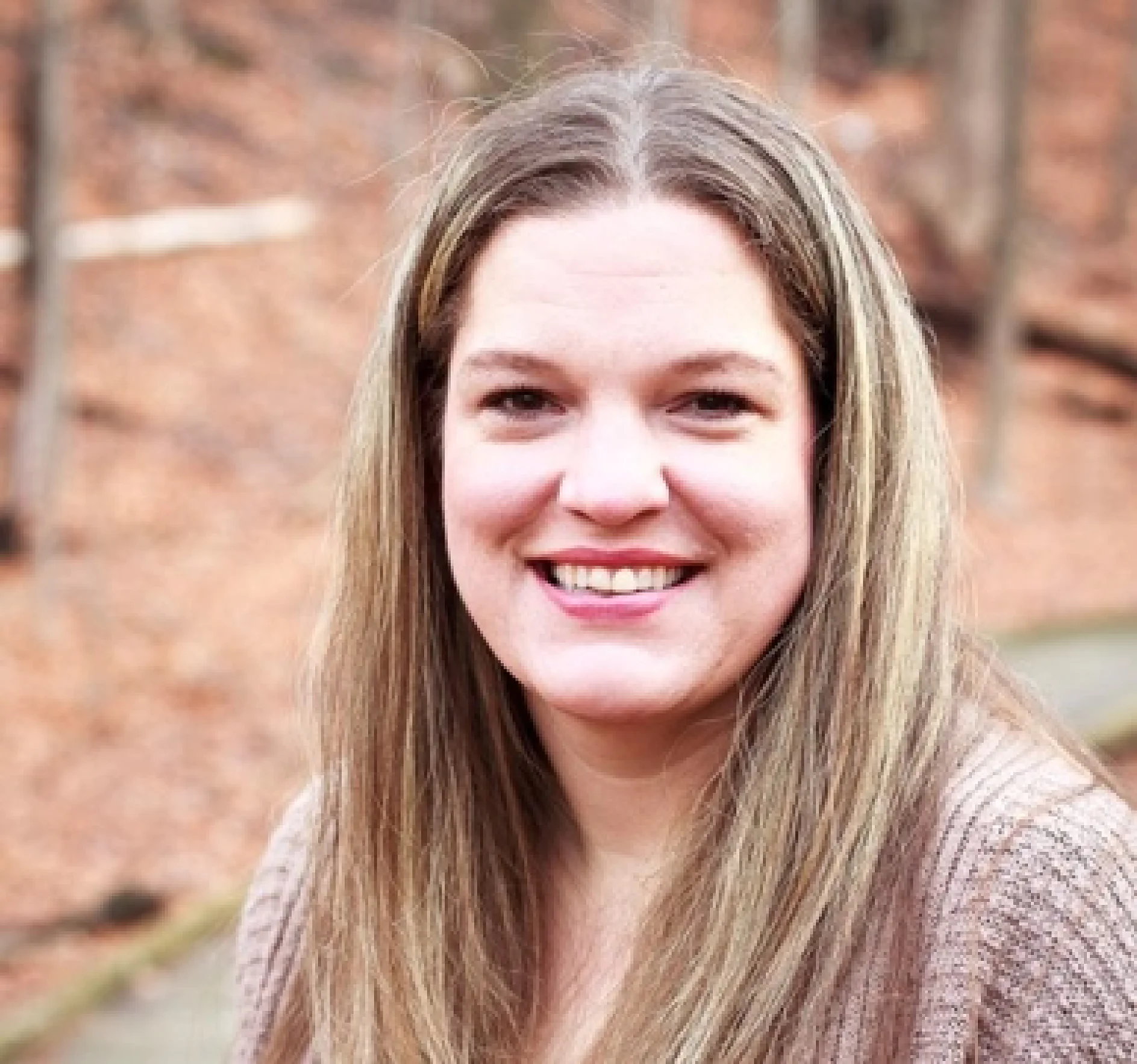 A woman with long, straight, light brown hair is smiling at the camera. She is wearing a beige sweater. The background appears to be an outdoor setting with blurred brown leaves and trees. (This text has been generated by AI)