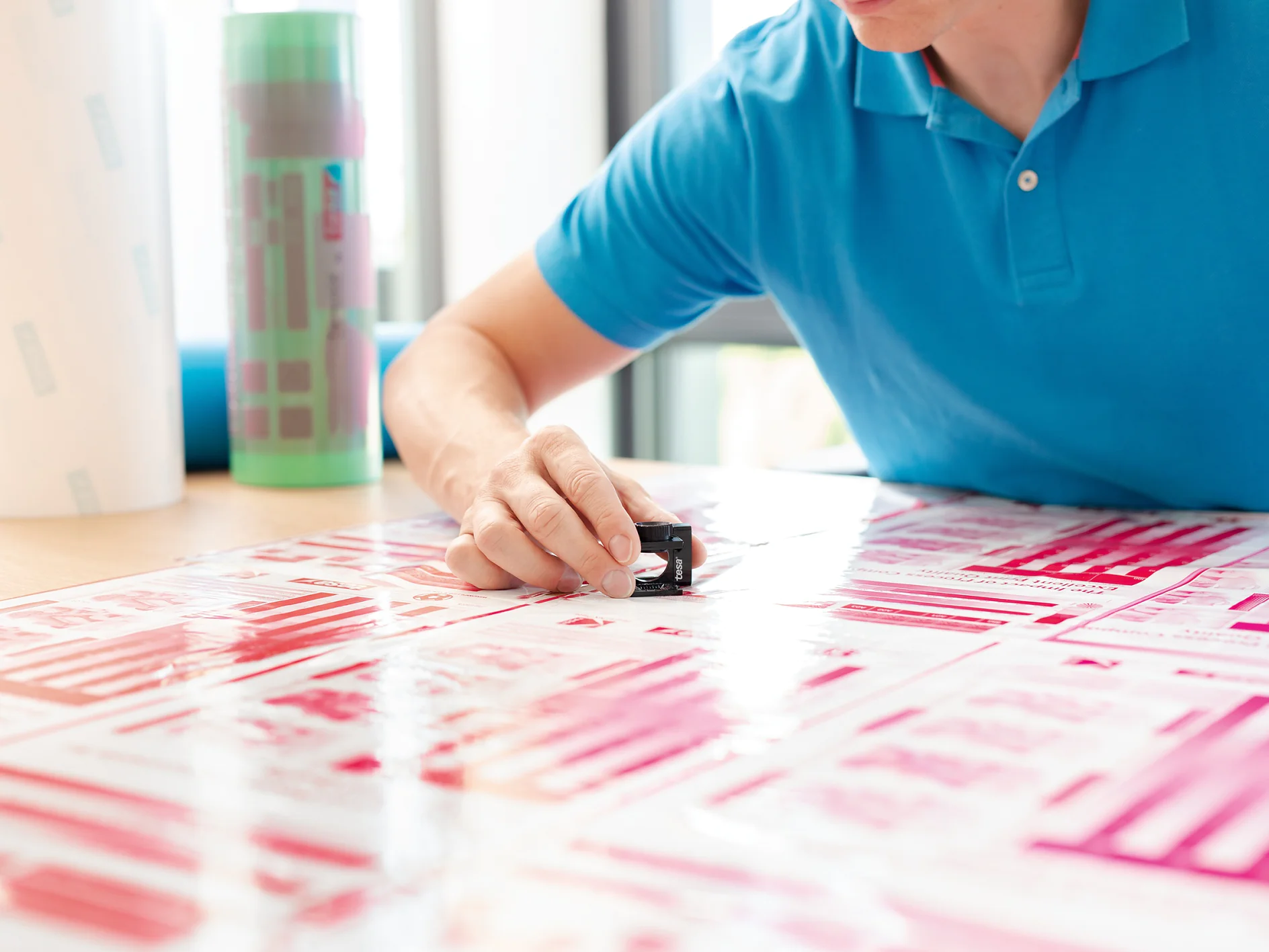 A person in a blue shirt examines printed materials with a magnifying tool on a table. The materials have various shades of pink and red, and there are rolls of tesa tape in the background. (This text has been generated by AI)