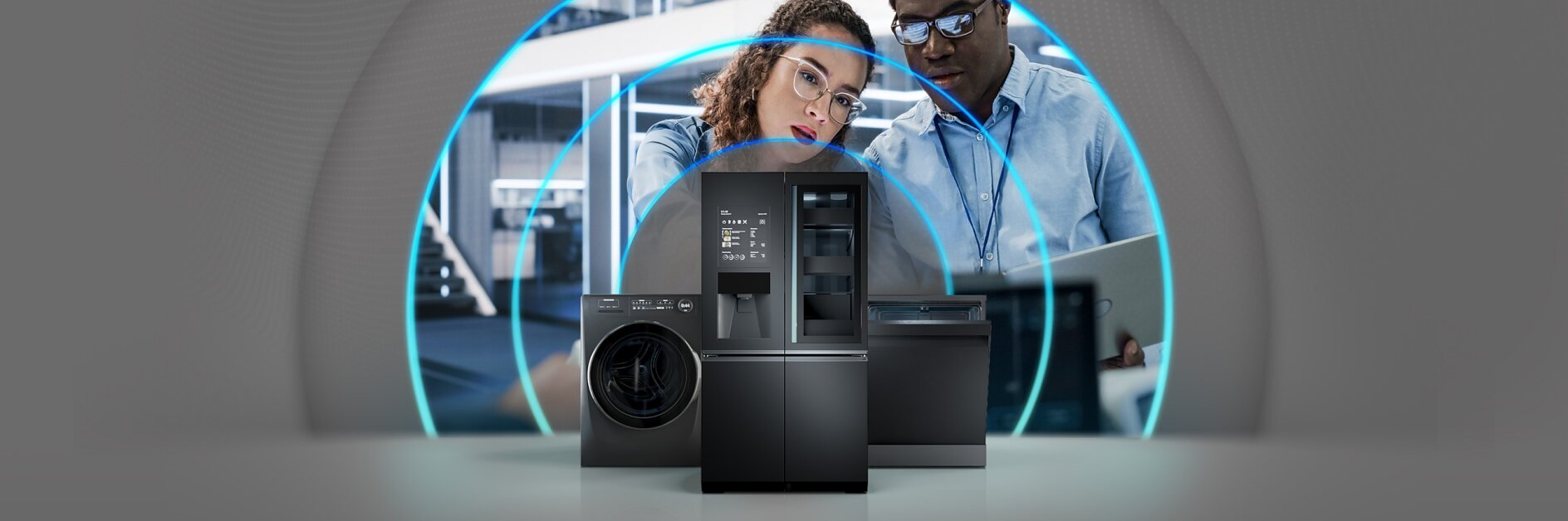A woman and a man looking at a laptop stand behind a black refrigerator, washing machine, and dishwasher. Blue circular lines surround them, suggesting technology or connectivity. The background shows a modern interior space. (This text has been generated by AI)